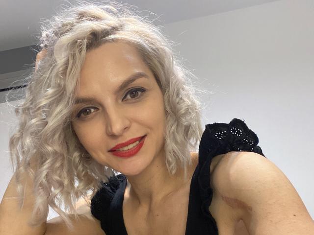 DouceAmelia with curly white hair, red lipstick, black dress, smiling, holding hair, light skin, brown eyes, visible arm bruise, selfie, white wall background, light grey ceiling.