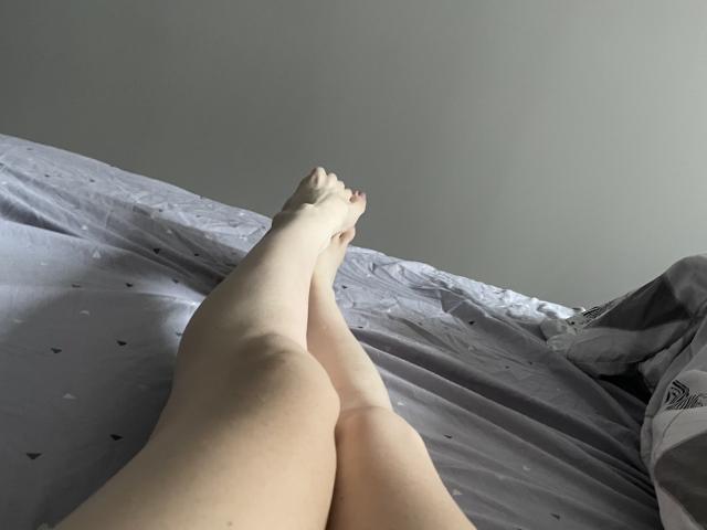 Las piernas pálidas de RozalinaPleasure, cruzadas a la altura de los tobillos, descansan sobre una cama gris con pequeños motivos de corazones. Sus pies están apuntando hacia arriba, mostrando una piel suave y tersa..