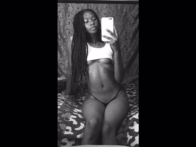 Black-and-white selfie of Keirysex with long braids, wearing a white crop top and black thong, sitting on a patterned blanket, holding a phone, showing her tits and ass.