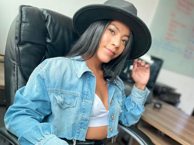Amberv with long black hair, wearing a black hat, blue denim jacket, and white bra, sitting in a black leather chair, hand near hat, slightly smiling.
