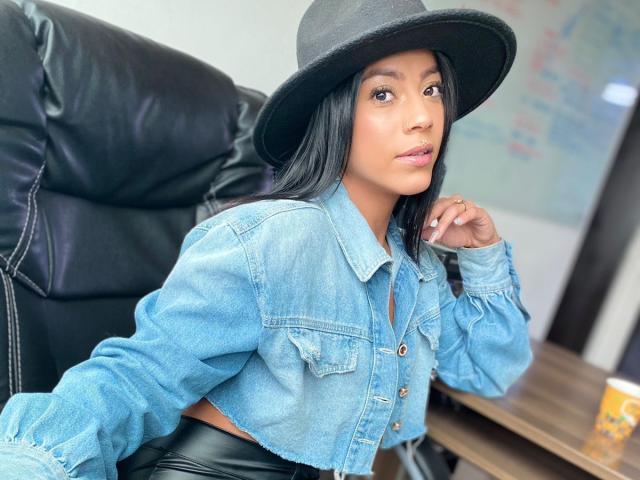 Amberv with long black hair, wearing a black hat, blue denim jacket, and black leather pants, sitting on a black leather chair, looking to the side, hand near mouth.