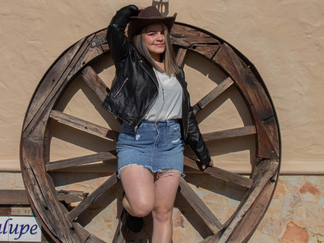 TokioParker stands against a wooden wagon wheel, wearing a brown hat, black leather jacket, white shirt, and frayed denim skirt. She's smiling, with one arm raised, showing her ass.
