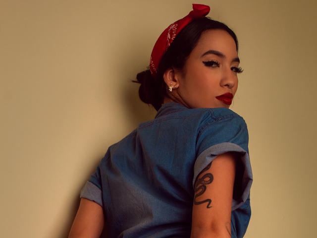 Kylie White, con cabello oscuro, pañuelo rojo, camisa vaquera azul, tatuaje en el brazo derecho, mirando por encima del hombro, lápiz labial rojo, delineador de ojos, aretes, pared beige, iluminación suave, sexy..