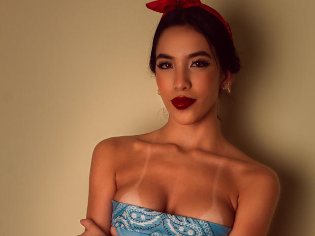 KylieWhite with tan lines on her tits, wearing a red bow in her dark hair, blue patterned strapless top, red lipstick, and small earrings, looking directly at the camera.