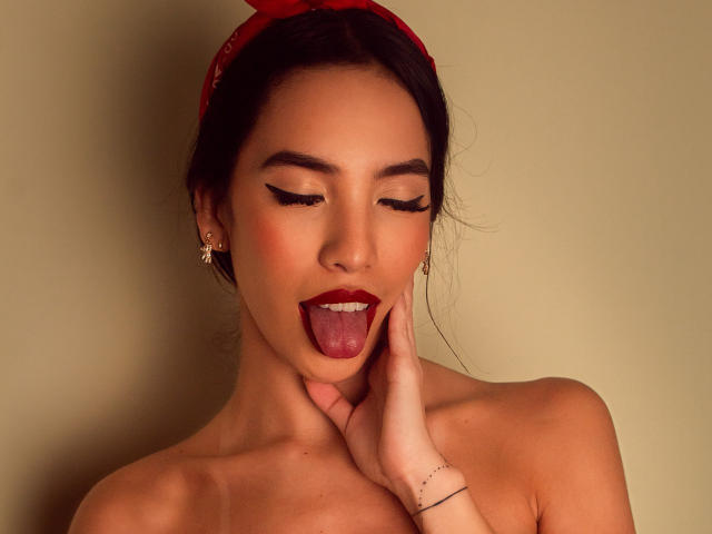 KylieWhite with dark hair, red headband, and earrings, tongue out, eyes closed, hand on neck, showing off red lipstick and sharp eyeliner, bare shoulders, cute and sexy.