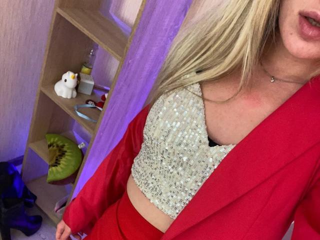 Alice, avec ses cheveux blonds, son blazer rouge et son crop top scintillant, dévoilant ses seins, portant un soutien-gorge noir en dessous, dans une pièce éclairée en violet et avec des étagères en bois..