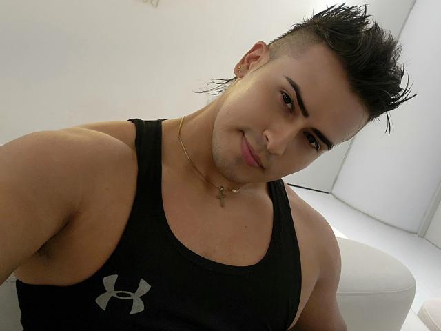 AngelLin has short, dark hair, a gold cross necklace, and a black Under Armour tank top, showing a confident, cute face with a slight smirk and a muscular, cute chest.