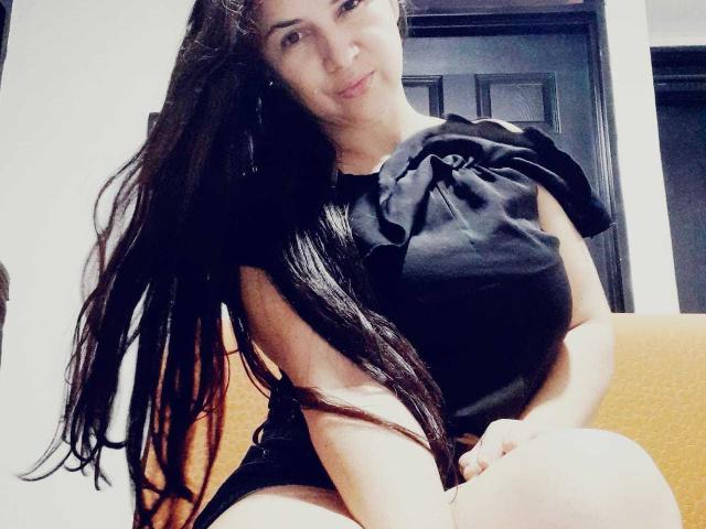 Brihana Charlot, com cabelos longos e pretos, vestindo uma blusa preta sem mangas, sentada em um sofá amarelo, sorrindo levemente, com as mãos apoiadas nos joelhos, exibindo sua nádega nua..