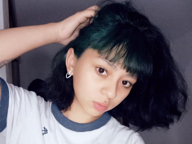 MefistoErica, avec des cheveux verts et noirs, des yeux marrons, un petit anneau de nez et une boucle d'oreille, portant une chemise blanche avec des détails bleus, une main dans les cheveux, regardant la caméra..