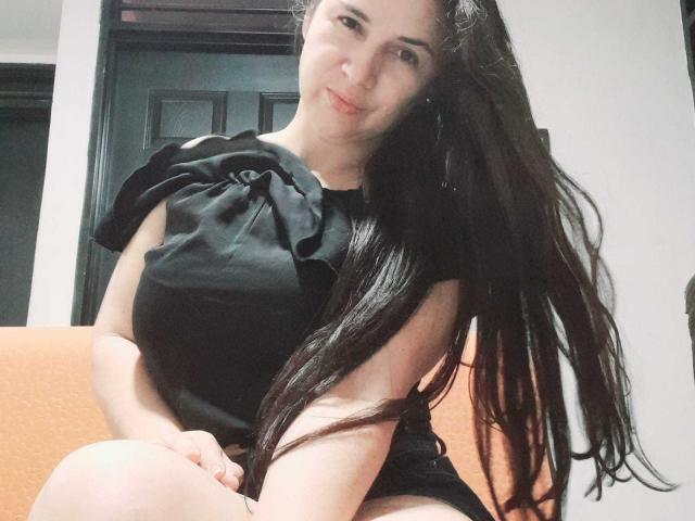 Brihana Charlot, com cabelos longos e pretos, sorrindo, vestindo um vestido preto com um grande laço, sentada em uma cadeira laranja, mostrando sua nádega nua, pele clara e rosto bonito..