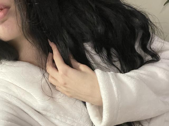 Les cheveux noirs et ondulés de RozalinaPleasure, sa robe blanche et moelleuse, et sa main pâle effleurant doucement ses cheveux, révélant une partie de ses lèvres roses et de ses doigts délicats..