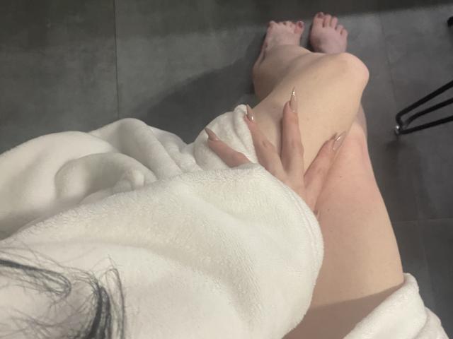 RozalinaPleasure a les jambes croisées, vêtue d'une serviette blanche, une main avec de longs ongles brillants reposant sur sa cuisse, les pieds nus sur des carreaux gris, une jambe de chaise noire en arrière-plan..