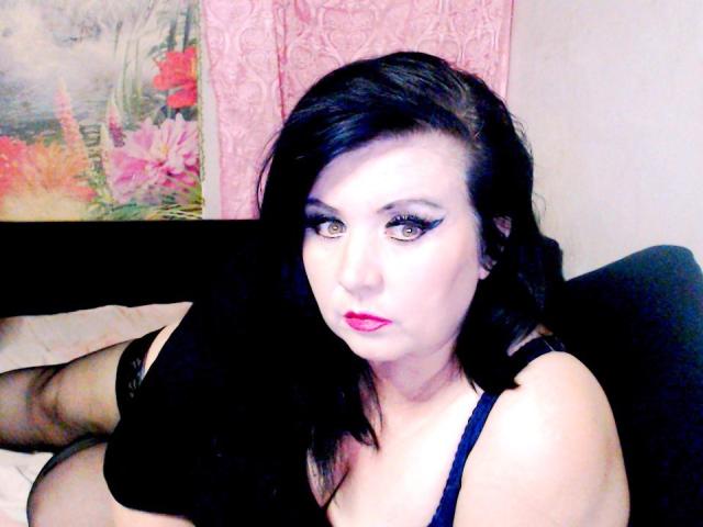 KamilaDream with black hair, pale skin, and pink lipstick, wearing a black top, looking at the camera with heavy black eyeliner, sitting on a bed, showing her tits.