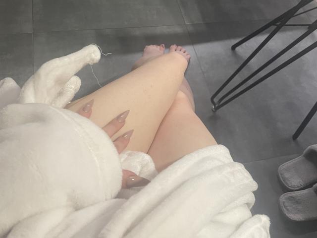 As pernas de RozalinaPleasure, vestindo um roupão branco e fofo, revelam a pele clara e unhas rosa nude, com os pés cruzados, perto de chinelos cinzas e pernas de uma cadeira preta. A vulva dela está parcialmente visível..