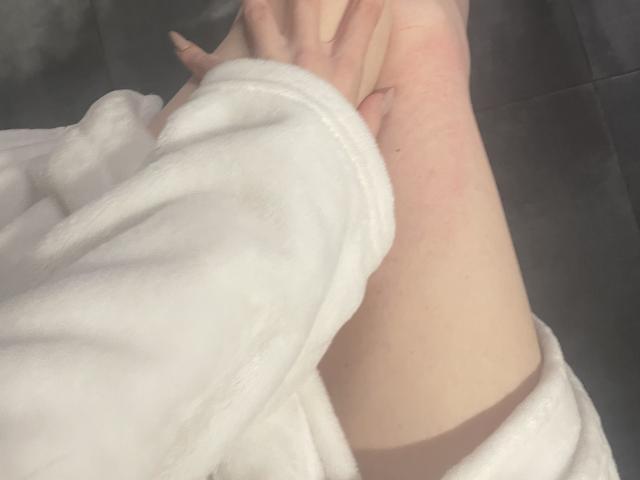 El brazo pálido de RozalinaPleasure, vestido con una túnica blanca, con una mano apoyada suavemente en su rodilla. La luz natural y suave resalta su piel. La textura de su túnica es ligeramente esponjosa..