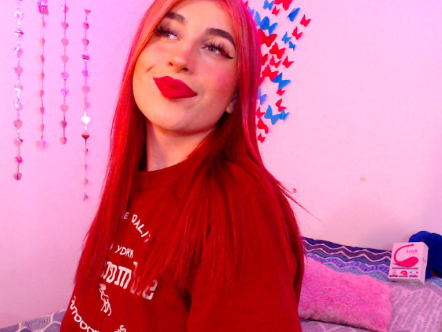 SammyViolet with red lipstick and long red hair, wearing a red shirt, smiling at the camera, pink and purple lighting, eyeliner, and a fluffy pillow in the background.