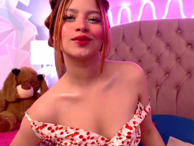 ShantalSummer has red lipstick, a nose ring, and is wearing a white dress with red heart patterns, showing her small tits and collarbone, with a cute brown teddy bear.