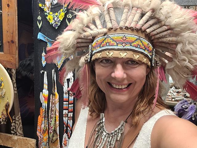 BarbaraBowie with fluffy white and pink-feathered headdress, smiling, wearing a beaded necklace, and a white sleeveless top. Her tits are visible under the top.