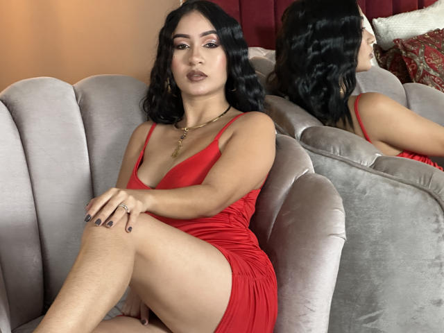 DevonPiper, com cabelo preto ondulado, vestindo um vestido vermelho, sentada em um sofá cinza, com as pernas cruzadas, mostrando sua vulva, reflexo no espelho, com uma expressão séria, usando um colar de ouro..