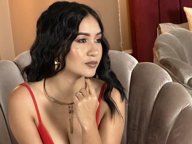 DevonPiper, com cabelos pretos ondulados, vestindo um vestido vermelho, brincos de ouro e um colar, tem a mão no queixo, exibindo seus seios e um anel no dedo..