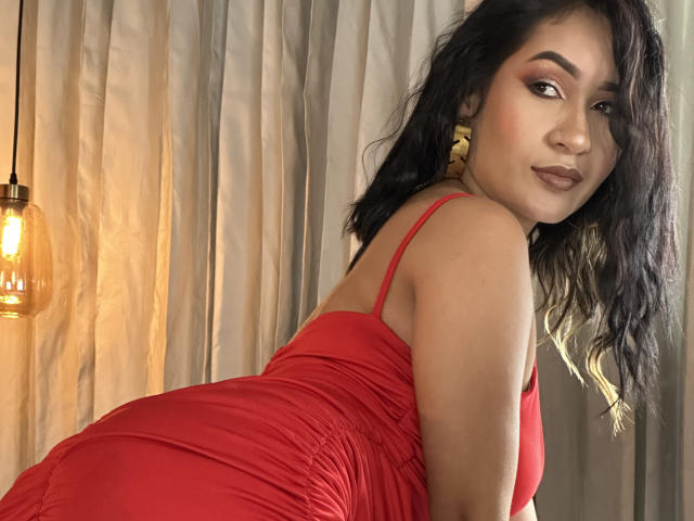 DevonPiper, vestindo um vestido vermelho, olhando por cima do ombro, exibindo suas nádegas, com cabelo preto ondulado, brincos de ouro e uma expressão sedutora. Luz quente de uma lâmpada pendurada..