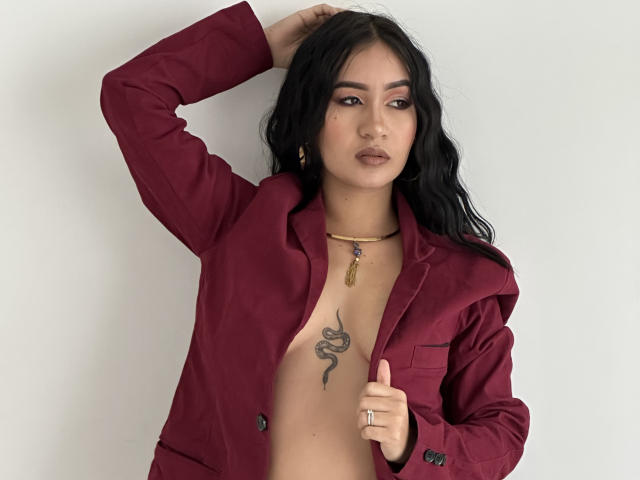 DevonPiper, com cabelo escuro, vestindo uma jaqueta marrom desabotoada, revelando uma tatuagem de cobra no peito, com a mão no cabelo, um anel no dedo, um colar de ouro, olhando para a câmera..