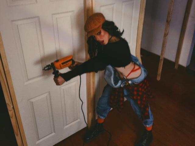 LucyVendeta, avec ses cheveux noirs bouclés, portant une casquette marron, un crop top noir, une chemise rouge à carreaux, un jean bleu et des chaussettes orange, utilisant une perceuse sur une porte blanche..