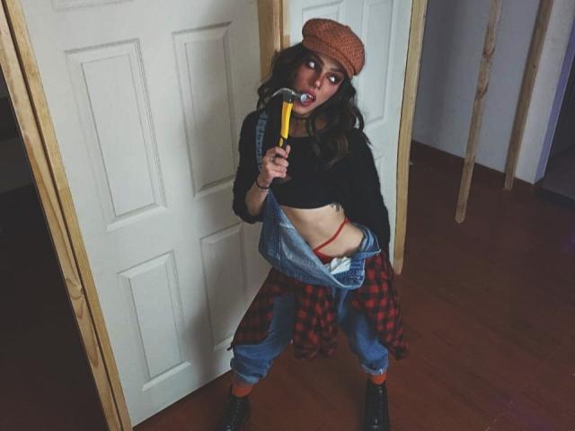 LucyVendeta está em uma porta, segurando um picolé amarelo, vestindo uma camisa xadrez vermelha, jeans azuis, uma blusa preta e um chapéu laranja, com uma calcinha vermelha visível, e botas pretas..