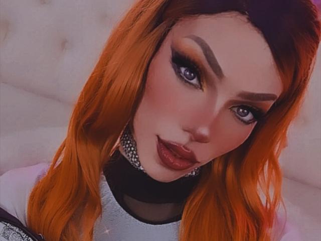 LucyVendeta a les cheveux d'un orange vif, un trait d'eye-liner audacieux et un rouge à lèvres violet foncé. Elle porte un collier noir et un haut scintillant. Ses fesses ne sont pas visibles..