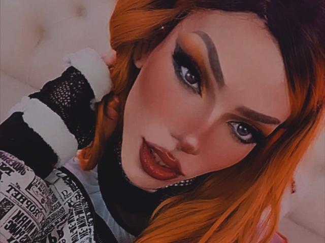 LucyVendeta tem cabelo laranja, sobrancelhas marcadas, delineador intenso e batom vermelho. Ela está vestindo uma blusa com estampa preta e branca, olhando para cima com a boca ligeiramente entreaberta e brilhante..