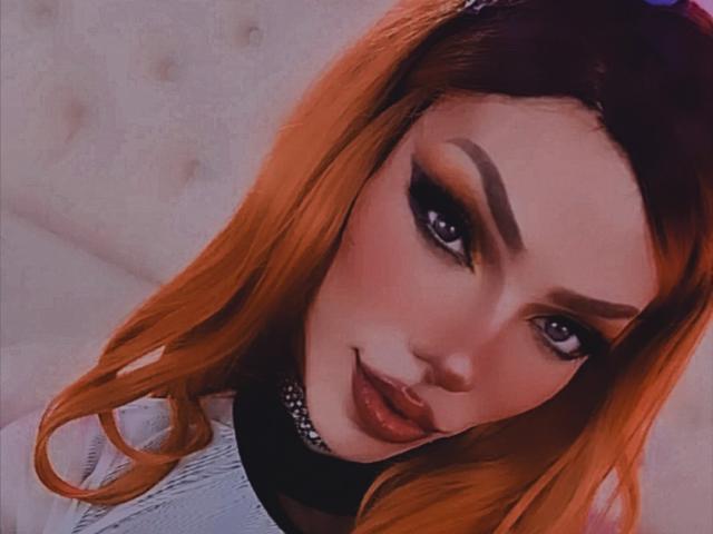 LucyVendeta, con cabello naranja, delineador de ojos marcado y lápiz labial rosa, lleva un collar y mira hacia arriba con una ligera sonrisa. Sus senos no son visibles en la imagen..