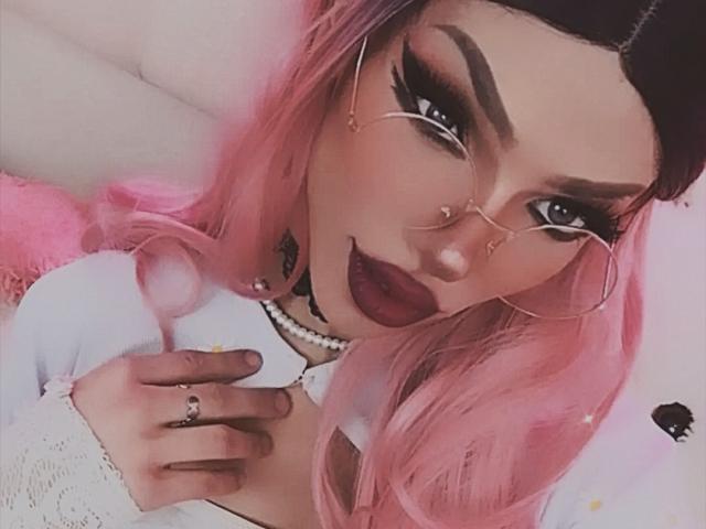 LucyVendeta, con cabello rosa, lápiz labial oscuro y aros de nariz, lleva gafas y una blusa blanca, sosteniéndose el pecho. Sus senos son pequeños y está mirando a la cámara..