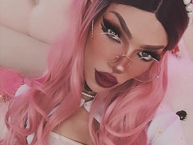 LucyVendeta ha i capelli rosa, il rossetto scuro e indossa occhiali con anelli al naso. Le sue sopracciglia folte e le lunghe ciglia sono molto evidenti. Sta guardando la fotocamera con un leggero sorriso..