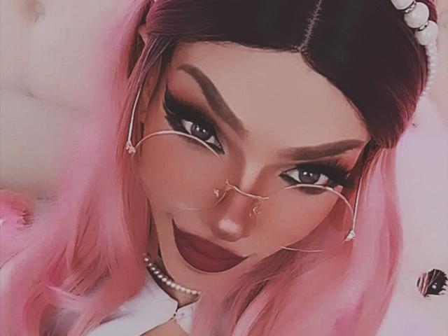 LucyVendeta, avec des cheveux roses, du rouge à lèvres foncé et du fard à paupières, portant des lunettes avec des chaînes et un collier de perles, souriant à la caméra. Seins partiellement visibles, fond blanc..