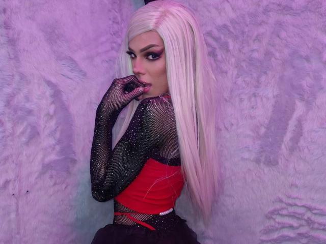 LucyVendeta, con cabello rubio largo, guantes de malla negra, una blusa roja y una falda negra, mordiéndose el dedo, mirando hacia atrás con ojos seductores. Su trasero es parcialmente visible..