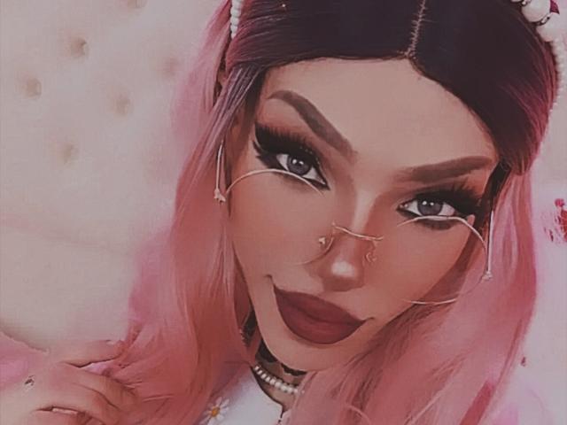 LucyVendeta, con cabello rosa, lápiz labial oscuro y pestañas gruesas, lleva un piercing en la nariz y gafas con cadena, sonriendo a la cámara, mostrando su rostro atractivo y su trasero lindo..