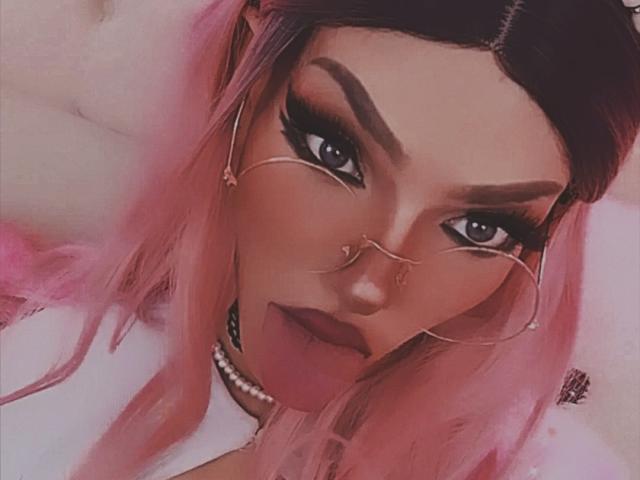 LucyVendeta, con i capelli rosa, le sopracciglia scure, gli orecchini a cerchio, il piercing al naso, gli occhiali, il rossetto scuro e la collana di perle. I suoi occhi guardano direttamente verso la fotocamera..