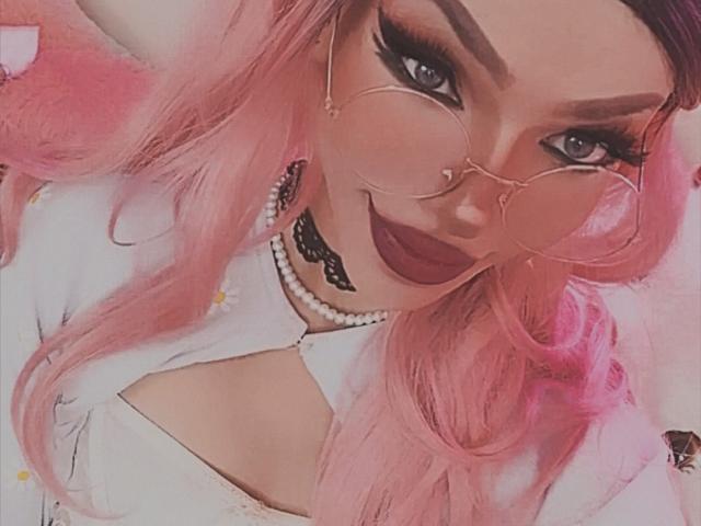 LucyVendeta, con cabello rosa, gafas redondas, lápiz labial oscuro, collar de perlas, collar de encaje negro y una blusa blanca, mostrando su rostro atractivo y su maquillaje impecable..