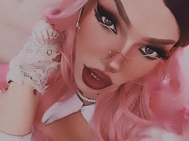 LucyVendeta, con i capelli rosa, ciglia folte, rossetto scuro, occhiali dorati senza lenti, collana di perle e top di pizzo bianco, che mostra il seno e i glutei in un selfie..