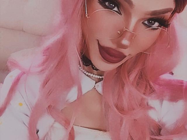 LucyVendeta, con cabello rosa, lápiz labial oscuro, piercing en la nariz y gafas, vestida con una camisa blanca y un collar de perlas, mostrando sus senos. Delicada, linda y sexy..