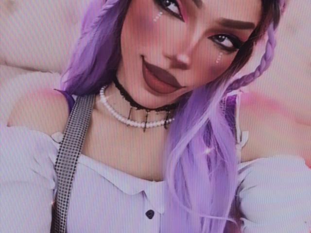LucyVendeta a les cheveux violets, un maquillage foncé et un collier de perles. Elle porte une chemise blanche à une épaule avec des bretelles grises. Ses seins sont petits et ses fesses ne sont pas visibles..