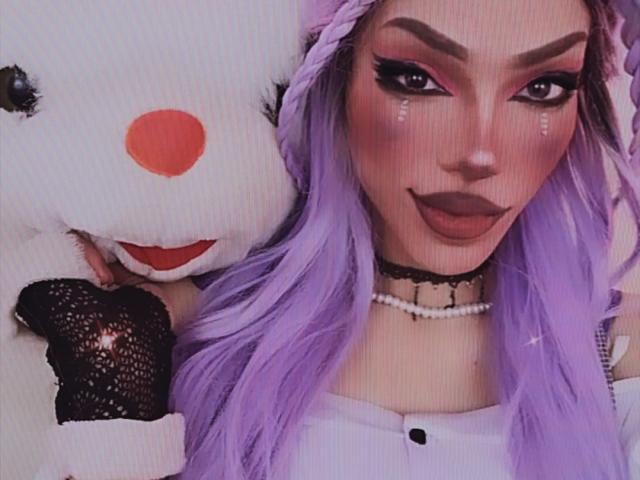 LucyVendeta, avec ses cheveux violets, son collier noir et son haut blanc, tenant un jouet en peluche blanc avec un nez rouge et une gante noire, et ses seins sont visibles..