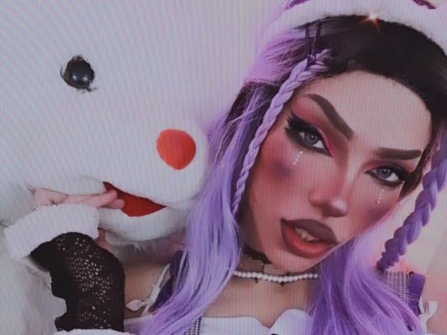 LucyVendeta, con i capelli viola, trecce e un cappello bianco, che tiene una pecora di peluche bianca con il naso rosso, indossa guanti di pizzo nero e una collana di perle, e appare sensuale..