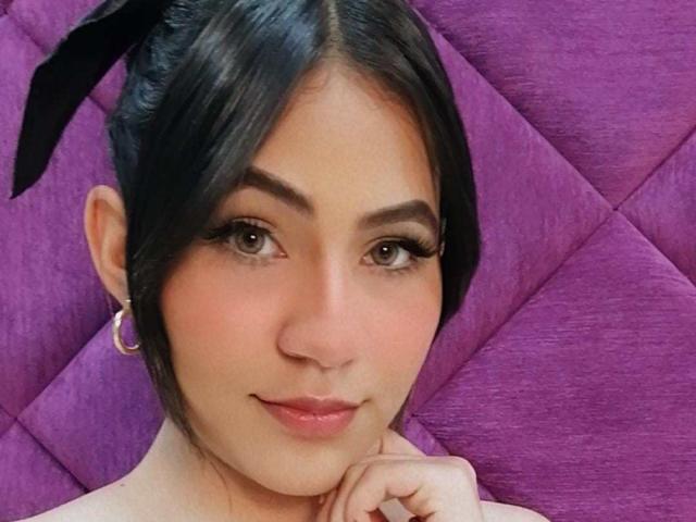 KataMillerr has black hair in a ponytail, brown eyes, thick eyebrows, and is wearing a gold hoop earring, with a pinkish nose and a slight smile, resting her chin.