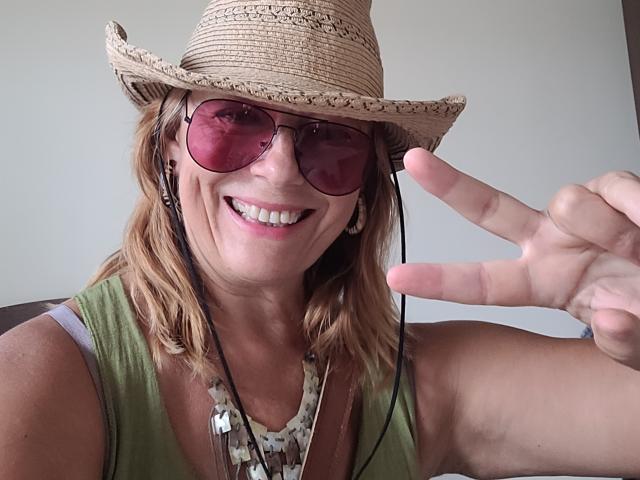 BarbaraBowie wearing a straw hat, pink sunglasses, green tank top, smiling, showing a peace sign, with blonde hair, necklace, and earbuds. Her tits are slightly visible.