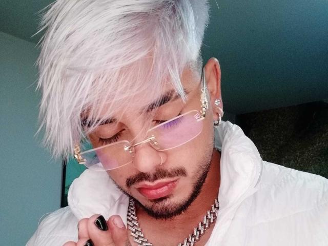 VisiI heeft wit, stekelig haar, roze brillen, een snor en een baardje, draagt een witte hoodie met een zilveren ketting en kijkt naar beneden met een zwarte nagel..
