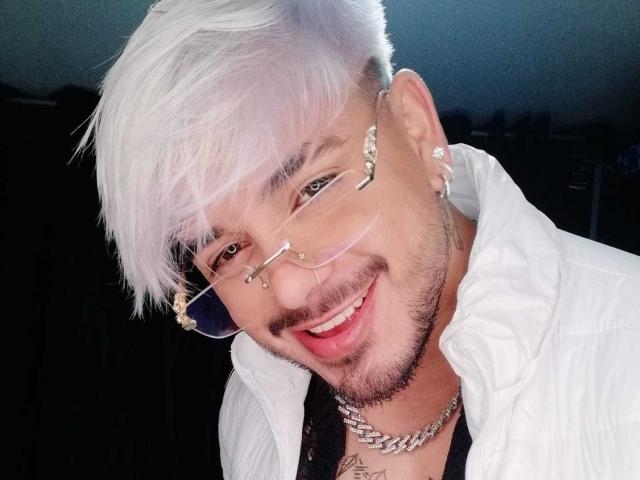 VisiI, met wit haar, neusringen en een bril, glimlachend met een baardje, draagt een witte jas en een ketting en heeft een tatoeage op zijn borst..
