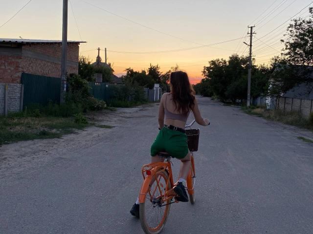 AurikaBriz riding an orange bike on a gravel road at sunset, wearing a white bra, green shorts, and black shoes, with long brown hair and a small black basket.