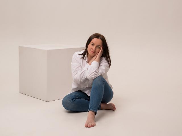 AbbyTaylorr is sitting barefoot with her hand on her cheek, wearing a white shirt and blue jeans, next to a white cube, looking cute.