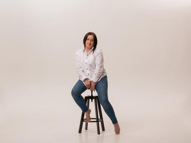 AbbyTaylorr sits on a stool, wearing a white shirt and blue jeans, with her tits slightly visible through the shirt, barefoot, and smiling.